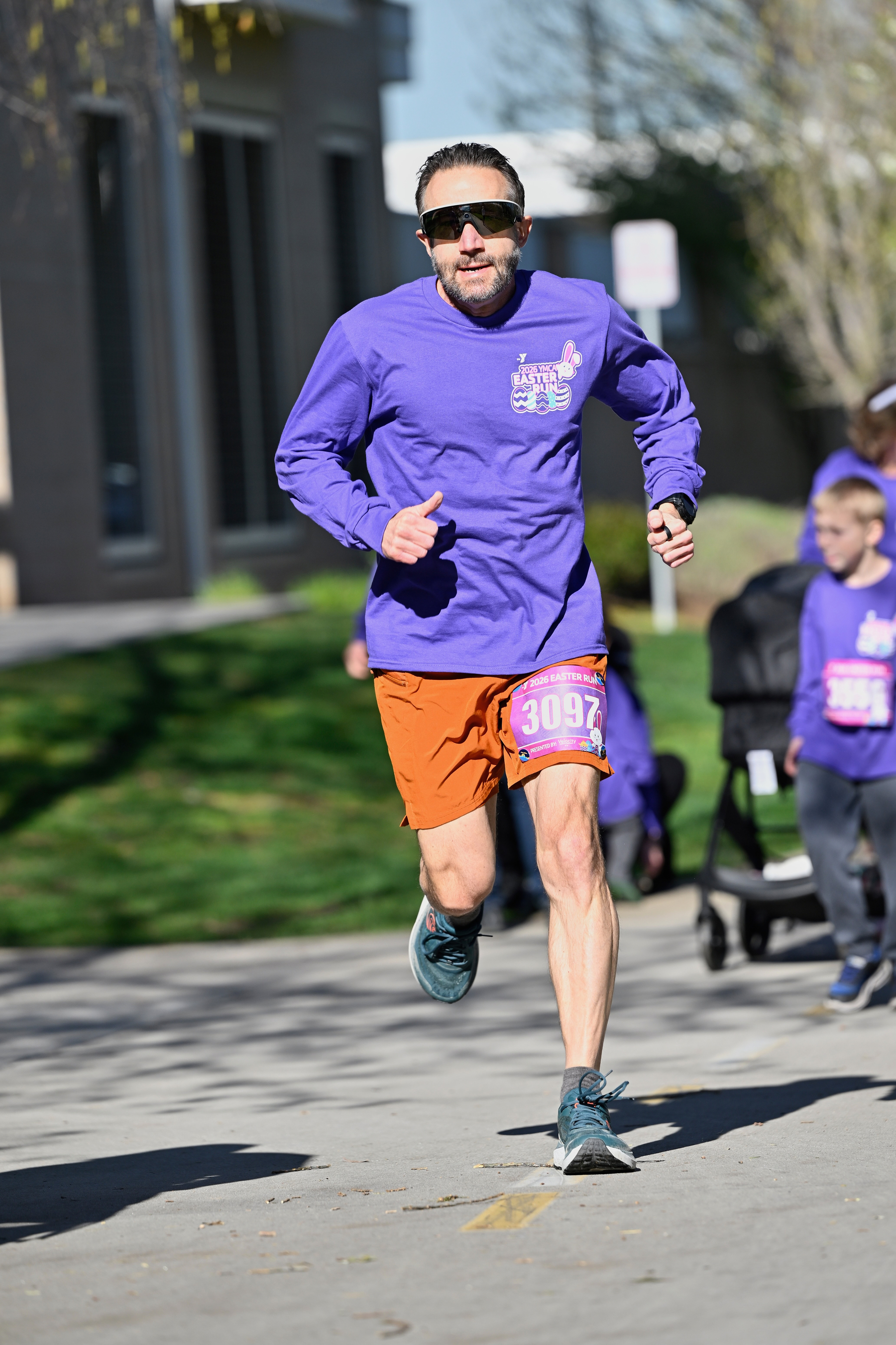 A man wearing a purple long-sleeve shirt and orange shorts is running on a paved path during a race event.